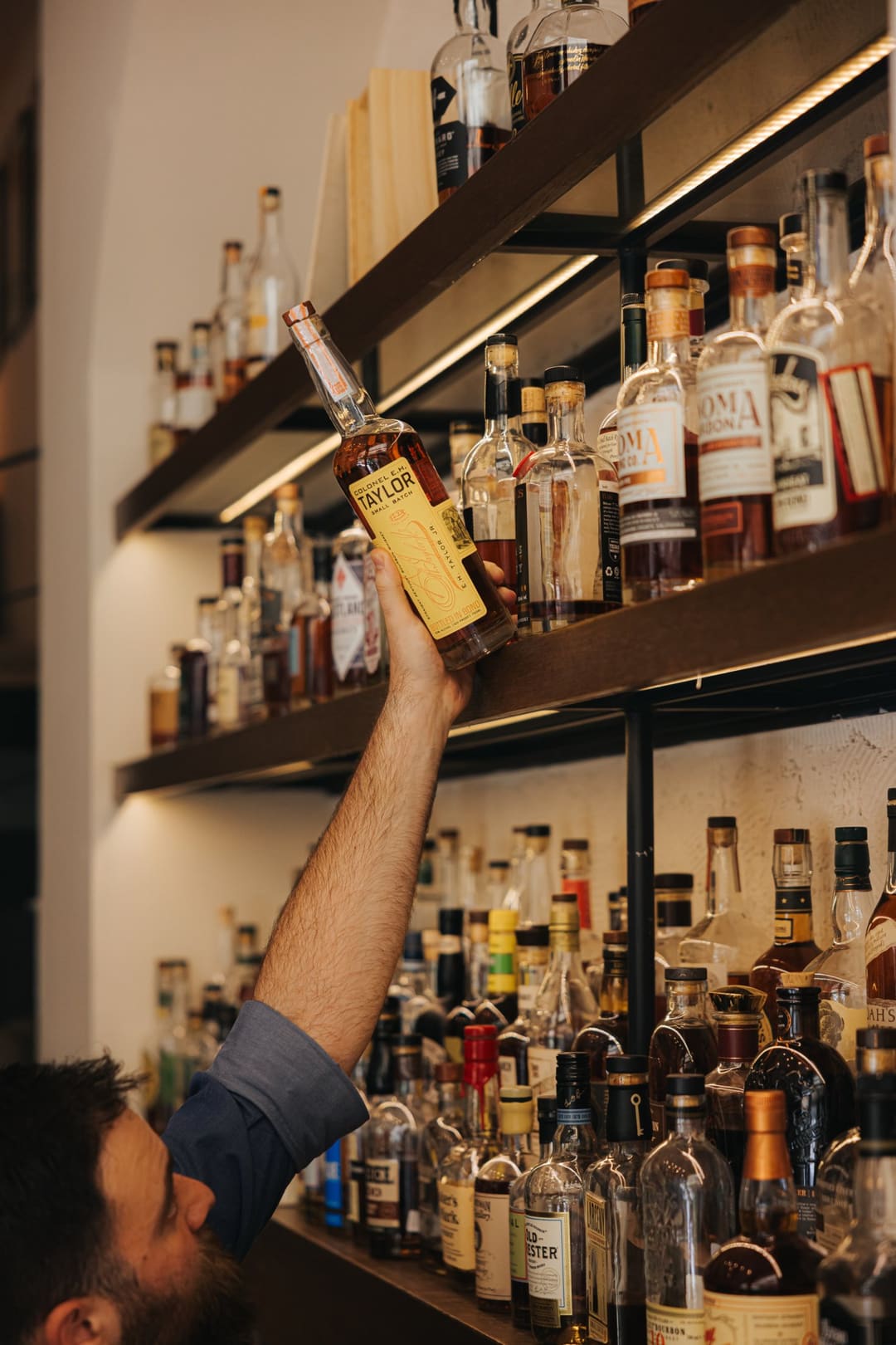 A bartender reaching for a bourbon bottle on the back bar shelves